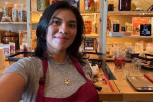 Kach in a fermentation workshop wearing an apron with shelves of jars and ingredients behind her.