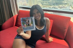 Kach smiling while sitting on a red couch, holding a 'Welcome Back Kach' card with her photo.